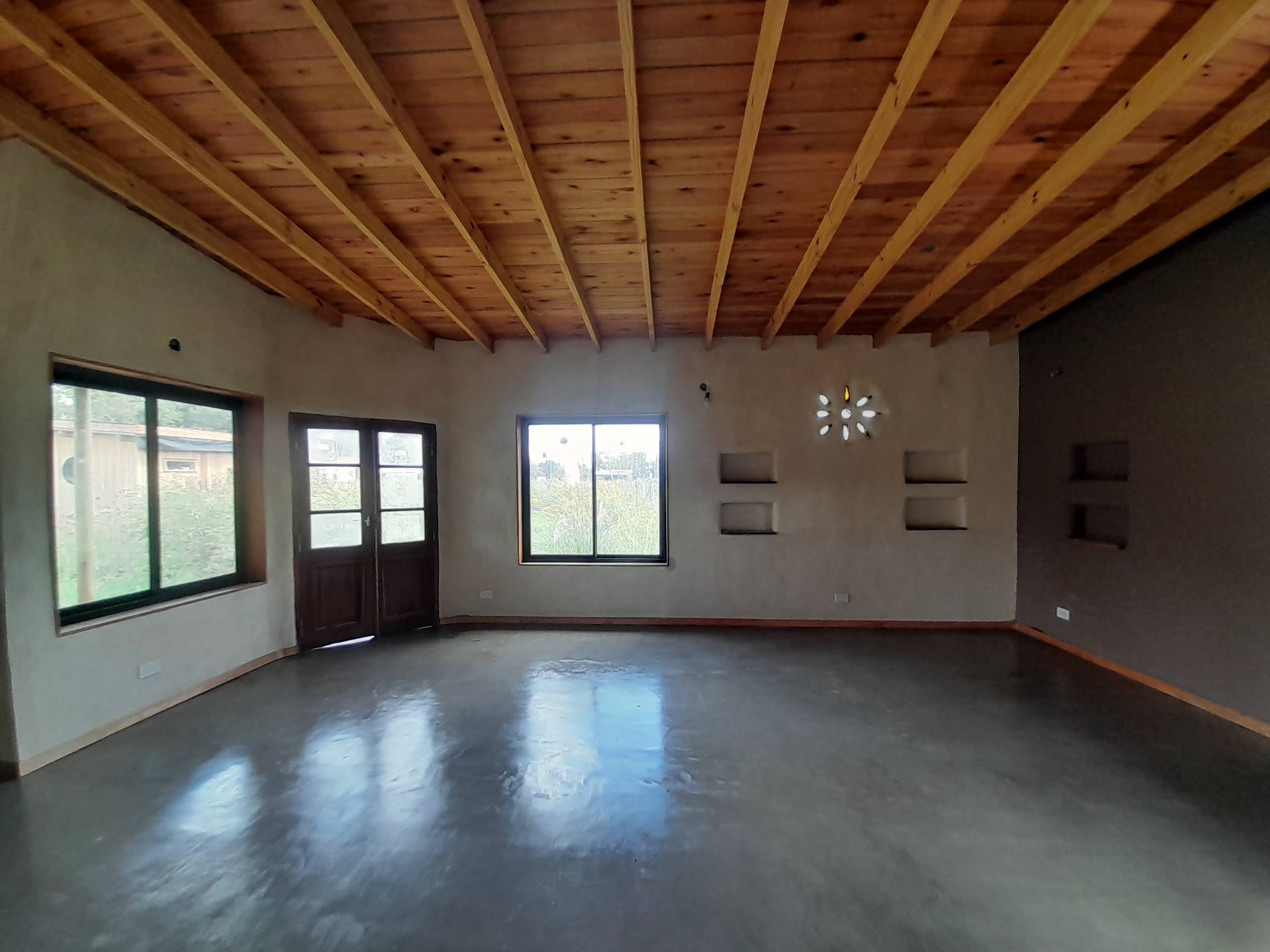 Un salón con paredes de color niebla, suelo brillante y techos de madera. Se ven ventanas principales y una puerta principal.