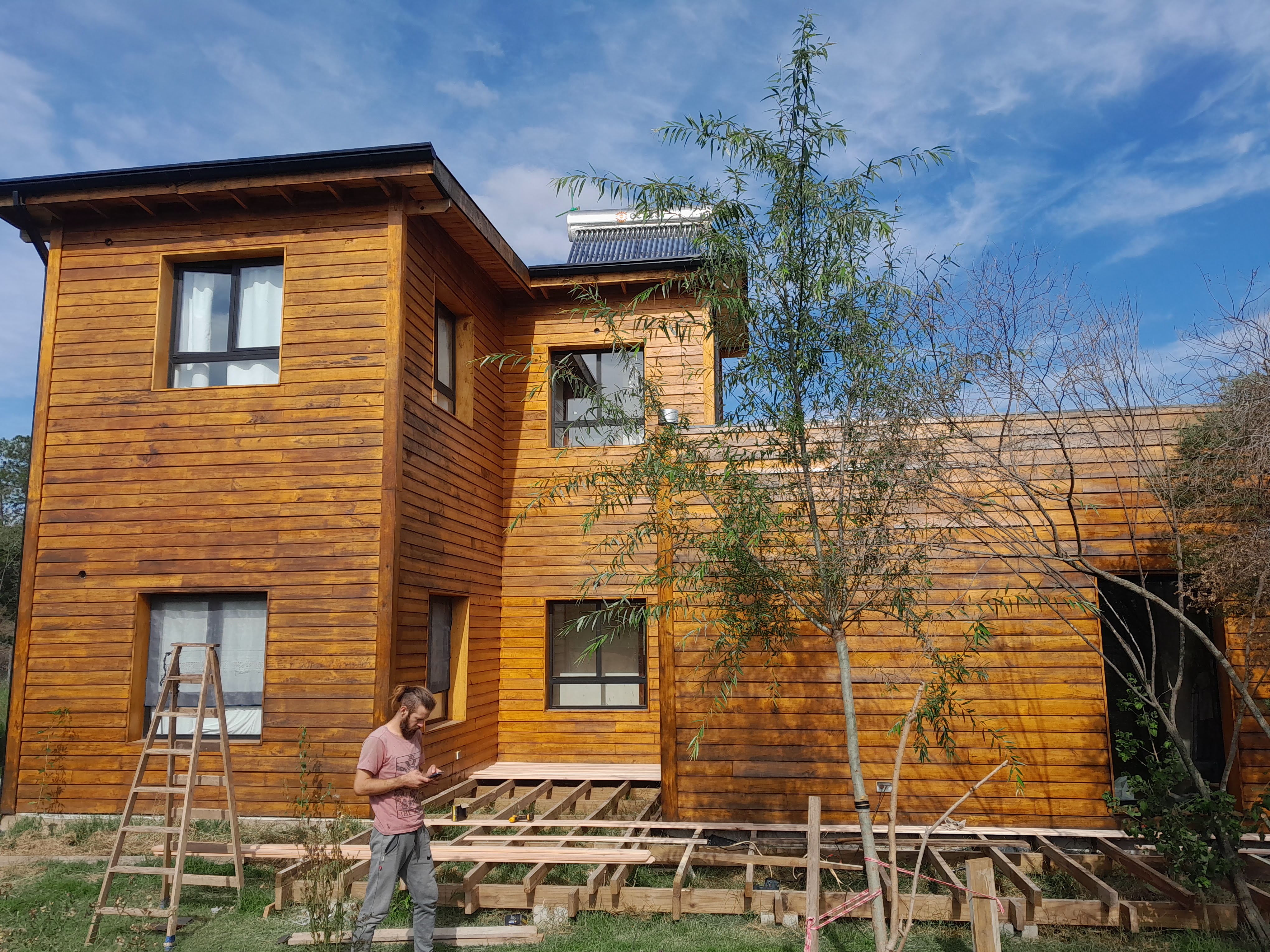 A man stands in front of a partially constructed wooden house with a brown exterior, surrounded by building materials and ...