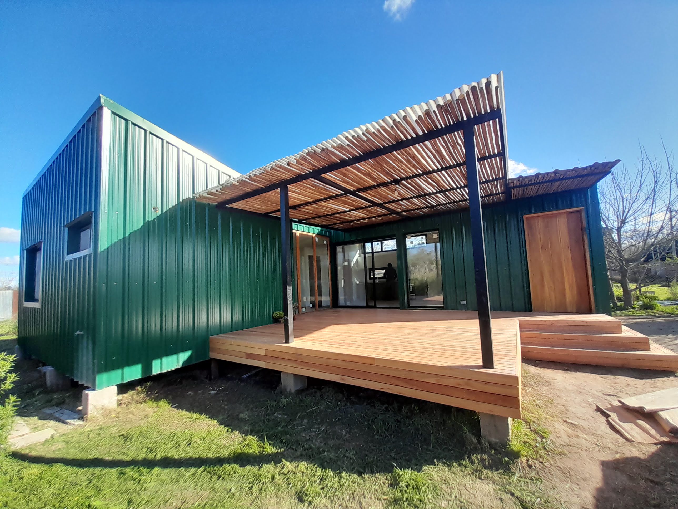 This image depicts a small, green metal-sided house with a wooden deck and a metal roof shade with a rustic touch. The str...